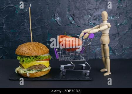 Figur eines kleinen Holzmanns, der einen Korb eines Supermarkts mit einer Tomate neben einem Hamburger schiebt. Stockfoto