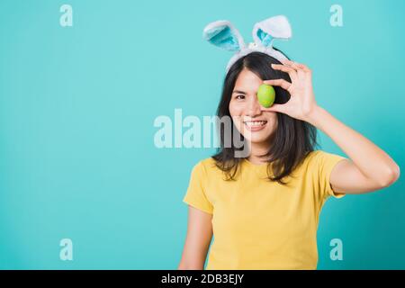 Portrait Asian schön fröhliche junge Frau lächelt weiße Zähne tragen gelbes T-Shirt, das mit Hünenohren steht und Ostereier in Augennähe auf blu hält Stockfoto