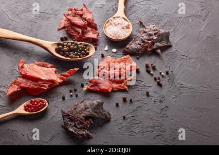 Ruckartig. Set aus verschiedenen Arten von getrocknetem gewürztem Fleisch, verschiedenen Pfefferkörnern und Salz auf Holzlöffeln auf dunkelgrauem Hintergrund. Draufsicht. Snack für Bier. Stockfoto