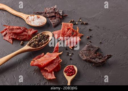 Ruckartig. Set aus verschiedenen Arten von getrocknetem gewürztem Fleisch, verschiedenen Pfefferkörnern und Salz auf Holzlöffeln auf dunkelgrauem Hintergrund. Draufsicht. Snack für Bier. Stockfoto