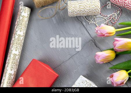Vorbereitung auf die Verpackung von Geschenken, auf dem Tisch aufgelegtes rotes und weißes Verpackungspapier, Fäden verschiedener Farben für Wickeln, Scheren und Tulpen Stockfoto
