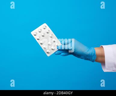 Weibliche Hand mit blauen sterilen Handschuhen hält eine Pille in einer Blase, Konzept für die medikamentöse Behandlung von Krankheiten, blauer Hintergrund Stockfoto