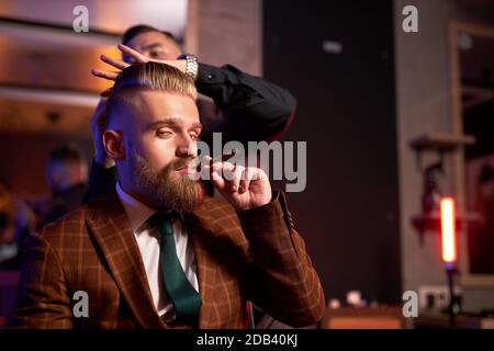 Junger kaukasischer Barbier, der die Haare seines kühlen reichen Klienten schneidet, der im Sessel sitzt und Zigarre raucht, Mann, der im Barberstuhl sitzt, er mag Haarschnitt. barbershop Konzept Stockfoto