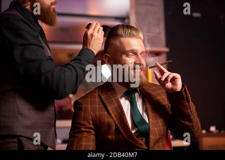 Junger kaukasischer Barbier, der die Haare seines kühlen reichen Klienten schneidet, der im Sessel sitzt und Zigarre raucht, Mann, der im Barberstuhl sitzt, er mag Haarschnitt. barbershop Konzept Stockfoto