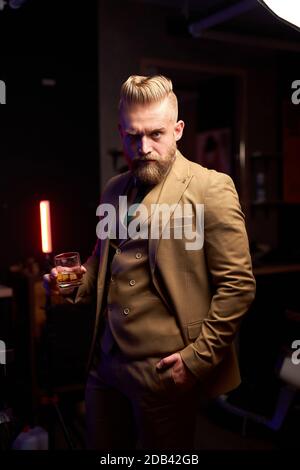 Der junge kaukasische bärtige Geschäftsmann trinkt drinnen im dunklen Raum ein Glas Whiskey und trägt einen formellen Anzug Stockfoto