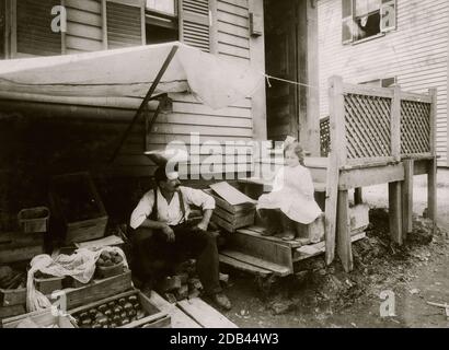 Haus und Obststand der Familie Cavicchio. 64 Bedford Street. Somerville, Mass. Sowohl die Mutter als auch Mary, 10 Jahre alt, arbeiteten den ganzen Tag hier mit dem Ruß der vorbeifahrenden Motoren, der so schnell abfiel, dass er abgeschüttelt werden musste. Vor dem Haus wird ein neuer Tunnel gegraben. Mutter und Maria pflegen den Fruchtstand beim Häkeln und die Arbeit wird über das Geländer und den Zaun in der Nähe geworfen. Ihre Kleider sind schmutzig und schmutzig. Stockfoto