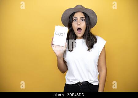 Schöne Frau trägt lässiges weißes T-Shirt und einen Hut stehen Über gelben Hintergrund erschrocken und überrascht halten Notebook mit Nein Word-Nachricht Stockfoto