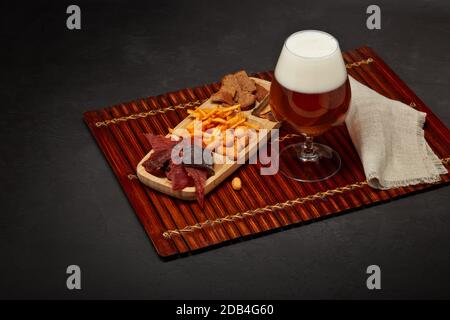 Glas mit Bier und verschiedene Snacks: Jerky, Kartoffelchips, gesalzene Nüsse, Stockfisch, Brotcracker auf braunem Holztablett auf dunkelgrauer Textur Stockfoto