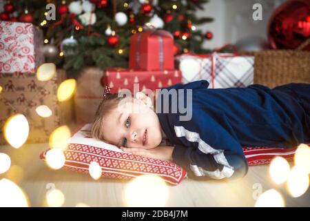 Ein kleines Mädchen in Heimkleidung liegt unter dem Weihnachtsbaum in der Nähe der Boxen mit Geschenken auf dem Kissen und Träumen. Lichter der Girlanden, die Nacht befo Stockfoto