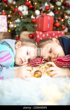 Ein kleines Mädchen in Heimkleidung liegt unter dem Weihnachtsbaum in der Nähe der Boxen mit Geschenken auf dem Kissen und Träumen. Lichter der Girlanden, die Nacht befo Stockfoto