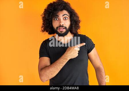 Junger arabischer bärtiger Rüde mit trendigen Haarspitzen mit Zeigefinger zur Seite, zeigen etwas, lockiger Kerl in Casual Wear isoliert über orangenen Hintergrund Stockfoto