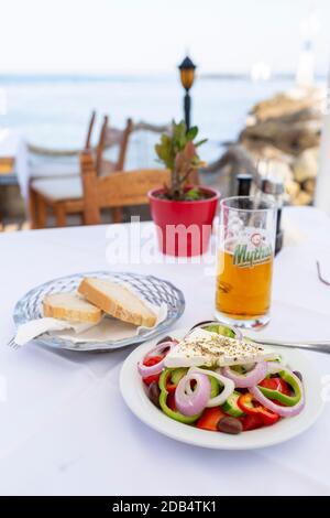 Griechischer Salat, lokales Bier und Brot, auf einem Tisch in einer Taverne am Meer in Georgioupoli, Kreta, Griechenland Stockfoto