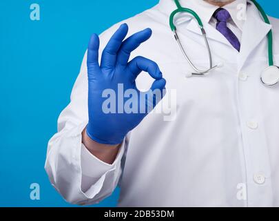 Männliche Ärztin in weißem Mantel, blaue Latexhandschuhe zeigen mit der rechten Hand eine gute Geste, das Konzept der Hoffnung, dass alles in Ordnung sein wird, blauer Backgrou Stockfoto