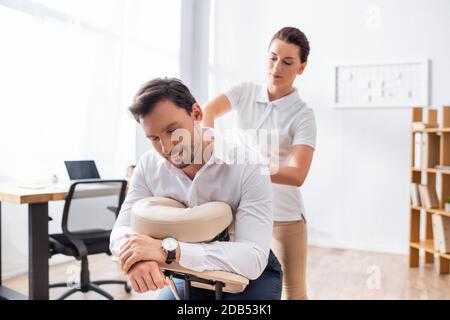 Weibliche Massage Therapeut massiert zurück von Geschäftsmann sitzen auf Massage Stuhl im Büro auf verschwommenem Hintergrund Stockfoto