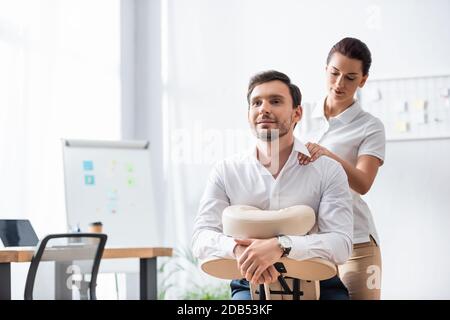 Masseuse Massage Schulter lächelnden Geschäftsmann sitzt auf Massagesessel Im Büro auf unscharfem Hintergrund Stockfoto