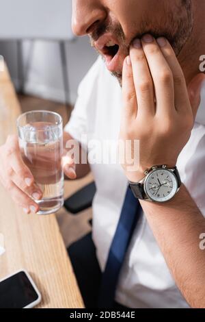 Beschnittene Ansicht des Geschäftsmannes mit der Hand auf der Wange, der an Zahnschmerzen leidet, während er ein Glas Wasser auf verschwommenem Hintergrund hält Stockfoto