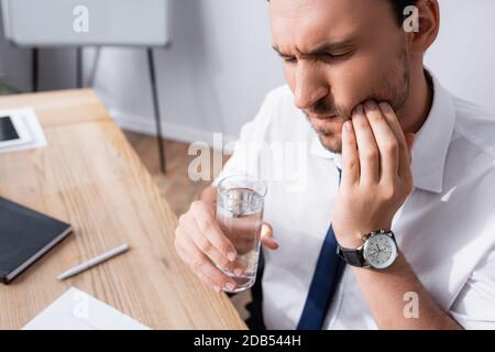 Geschäftsmann hält ein Glas Wasser und leidet an Zahnschmerzen, während er auf unscharfem Hintergrund am Arbeitsplatz sitzt Stockfoto