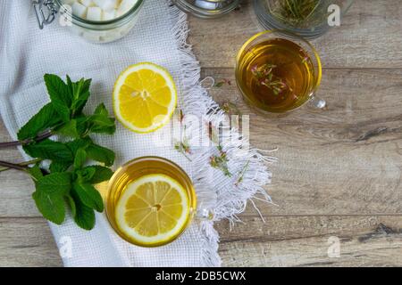 Glas transparente Tasse mit Minztee steht auf einem Holzständer, neben einer Zuckerdose mit Zuckerwürfeln, in Scheiben geschnittenen Zitronen- und Minzblättern. Stockfoto