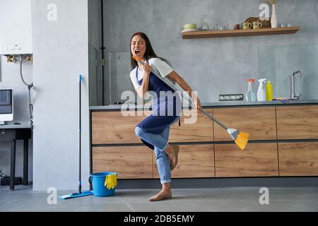 In voller Länge Schuss von fröhlichen jungen Frau, Putzfrau vorgibt, ein Lied zu singen, hält Besen während der Reinigung des Bodens, Hausarbeit zu tun. Hausarbeit und Housekeeping Konzept Stockfoto