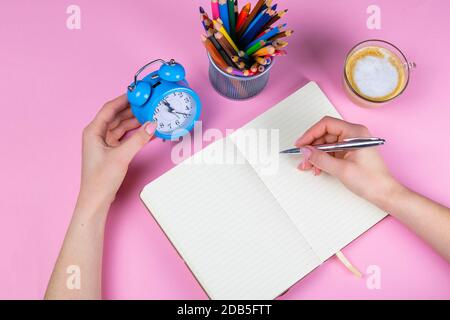 Tablette mit weißem Blatt, neben dem ist eine grüne Pflanze, ein Glas mit Bleistiften. Die Hand hält einen Stift und schreibt eine Uhr in eine andere Hand auf das Blatt. Stockfoto