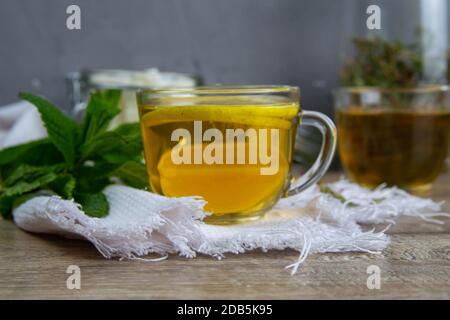 Glas transparente Tasse mit Minztee steht auf einem Holzständer, neben einer Zuckerdose mit Zuckerwürfeln, in Scheiben geschnittenen Zitronen- und Minzblättern. Stockfoto