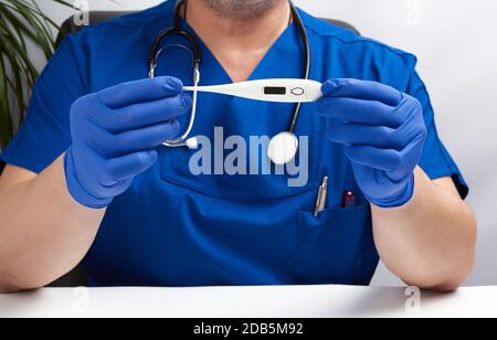 Doktor sitzt in blauer Uniform und Latexhandschuhe hält ein weißes elektronisches Thermometer, Arztpraxis Stockfoto