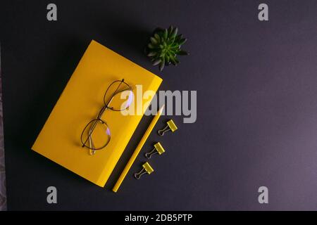 Gelber Bleistift, gelbes Notizbuch und Brille, Stifte, Clip-Papier auf schwarzem Hintergrund. Stockfoto