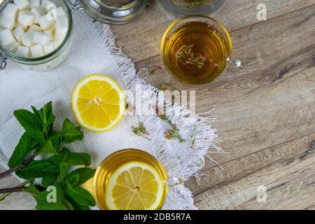 Glas transparente Tasse mit Minztee steht auf einem Holzständer, neben einer Zuckerdose mit Zuckerwürfeln, in Scheiben geschnittenen Zitronen- und Minzblättern. Stockfoto