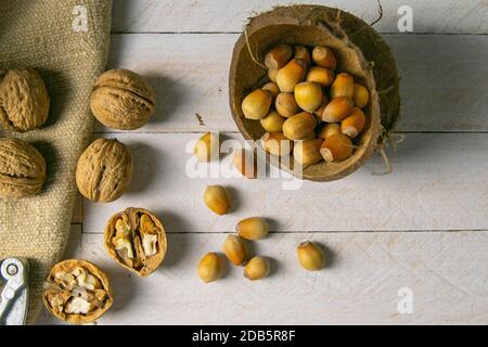 Walnüsse in einer Holzkiste neben einer Haselnuss in einer Kokosnussschale, Nüsse auf einem weißen Holztisch verstreut, auf dem auch der Nussknacker steht. Stockfoto