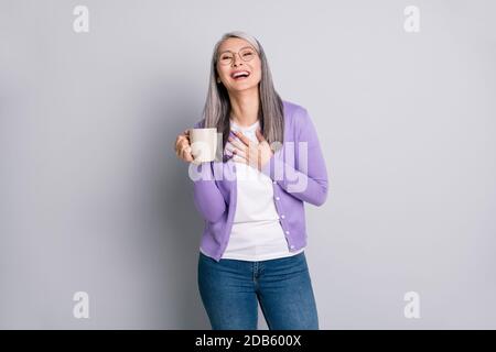 Foto Porträt einer Frau lachend hält Tasse in einer Hand Berührende Brust mit anderen tragen violetten Cardigan isoliert auf grau Farbiger Hintergrund Stockfoto