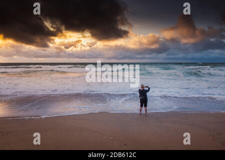 Ein Urlauber, der mit seinem Handy einen spektakulären Sonnenuntergang über Fistral Beach in Newquay in Cornwall fotografiert. Stockfoto