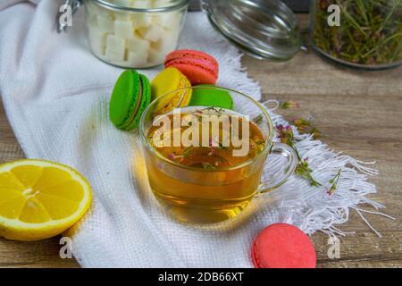 Glas transparente Tasse mit Minztee steht auf einem Holzständer, neben einer Zuckerdose mit Zuckerwürfeln, in Scheiben geschnittene Zitrone, Löffel und bunten Muffins. Stockfoto