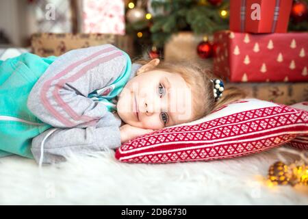 Ein kleines Mädchen in Heimkleidung liegt unter dem Weihnachtsbaum in der Nähe der Boxen mit Geschenken auf dem Kissen und Träumen. Lichter der Girlanden, die Nacht befo Stockfoto