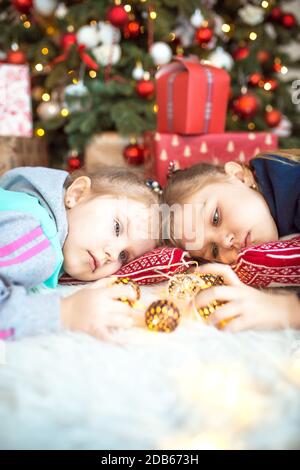 Ein kleines Mädchen in Heimkleidung liegt unter dem Weihnachtsbaum in der Nähe der Boxen mit Geschenken auf dem Kissen und Träumen. Lichter der Girlanden, die Nacht befo Stockfoto