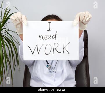 Junge Frau Doktor in einem weißen Mantel, sterile medizinische Handschuhe halten ein Plakat mit der Aufschrift "Ich brauche Arbeit", Begriff der Arbeitslosigkeit und Arbeitssuche Stockfoto