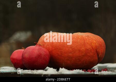 Das Stillleben von orangefarbenem Kürbis und roten Granatäpfeln liegt auf einer schneebedeckten Holzfläche im Freien vor dem Hintergrund eines Herbstgartens. Stockfoto
