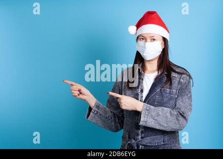 Eine asiatische Frau trägt Santa Hut und medizinische Schutzmaske gegen Virus, zeigt mit Zeigefinger auf leeren Raum Stockfoto