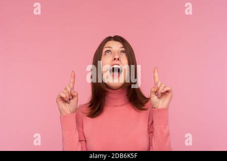 Extrem überrascht glückliche Frau mit braunen Haaren in rosa Pullover zeigt Finger nach oben auf leeren Raum, zufrieden mit Werbefläche. Innenstudio s Stockfoto