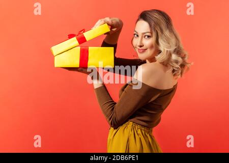 Seitenansicht schöne Frau Auspacken große gelbe Geschenkbox und Blick auf die Kamera mit angenehmen Lächeln, zufrieden mit Geschenk. Innenaufnahme des Studios isoliert Stockfoto