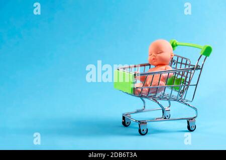 Puppe sitzt auf einem Einkaufswagen auf einem blauen Hintergrund. Eco, Leihmutterschaft, kaufen Sie ein Kind für Geld Stockfoto