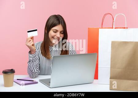 Glückliche aufgeregt Frau sitzt am Laptop und hält Kreditkarte, tun Online-Shopping, Papiertaschen liegen auf dem Schreibtisch, schnelle Lieferung Service. Innenstudio s Stockfoto