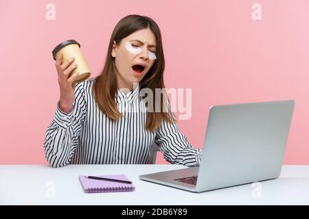 Müde schläfrig Frau freiberuflich gähnend Kaffee aus der Papiertasse trinken und arbeiten auf Laptop sitzen am Schreibtisch mit Augenklappe, arbeiten von zu Hause aus. Stehbolzen für den Innenbereich Stockfoto