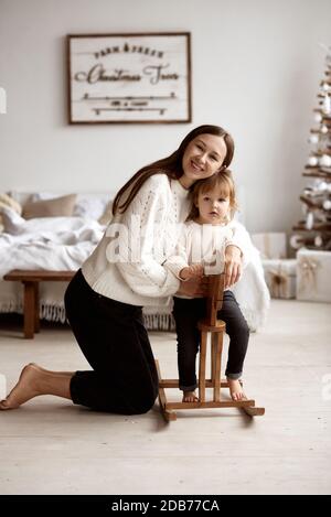 Junge liebevolle Mutter umarmt ihre Tochter, die auf einem hölzernen Schaukelpferd in einem hellen Raum sitzt. Stockfoto