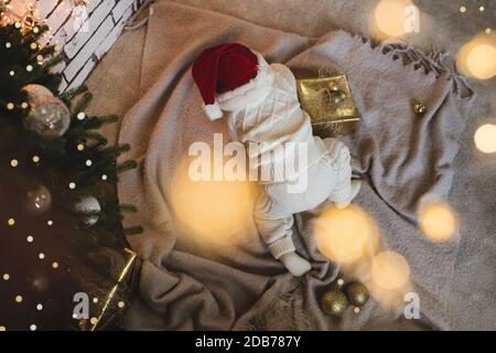 Baby unter 1 Jahr alt trägt weihnachtsmann Hut und Strickanzug kriechen unter Weihnachtsbaum im Zimmer über glühende Lichter aus der Nähe. Draufsicht. Kindheit Stockfoto