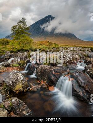 Malerischer Wasserfall am Fluss mit Buacheille Etive Mor im Hintergrund, umgeben von Morgennebel. Stockfoto