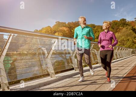 Bleiben Sie in Form. In voller Länge ein aktives, reifes Familienpaar in Sportswear, das sich beim Joggen an einem sonnigen Herbsttag anlächelt Stockfoto