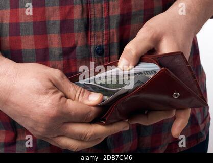 Mann in einem karierten roten Hemd hält eine braune Ledertasche voller einhundert US-Papier-Dollar-Banknoten, Begriff des Gehalts, Reichtum Stockfoto