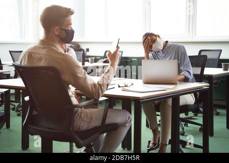 Junge frustrierte gemischte Rasse Frau trägt Schutzmaske berühren Kopf, während am Schreibtisch mit männlichen Kollegen sitzen, zusammen im Büro arbeiten Stockfoto