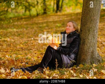 Ein schönes junges blondes Mädchen sitzt auf gefallenen Herbstblättern im Park und lehnt ihren Rücken gegen einen Baumstamm. Das Mädchen hält gelbe Ahornblätter Stockfoto