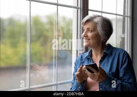 Lächelnd verträumt reife Frau hält Telefon, Blick aus dem Fenster Stockfoto